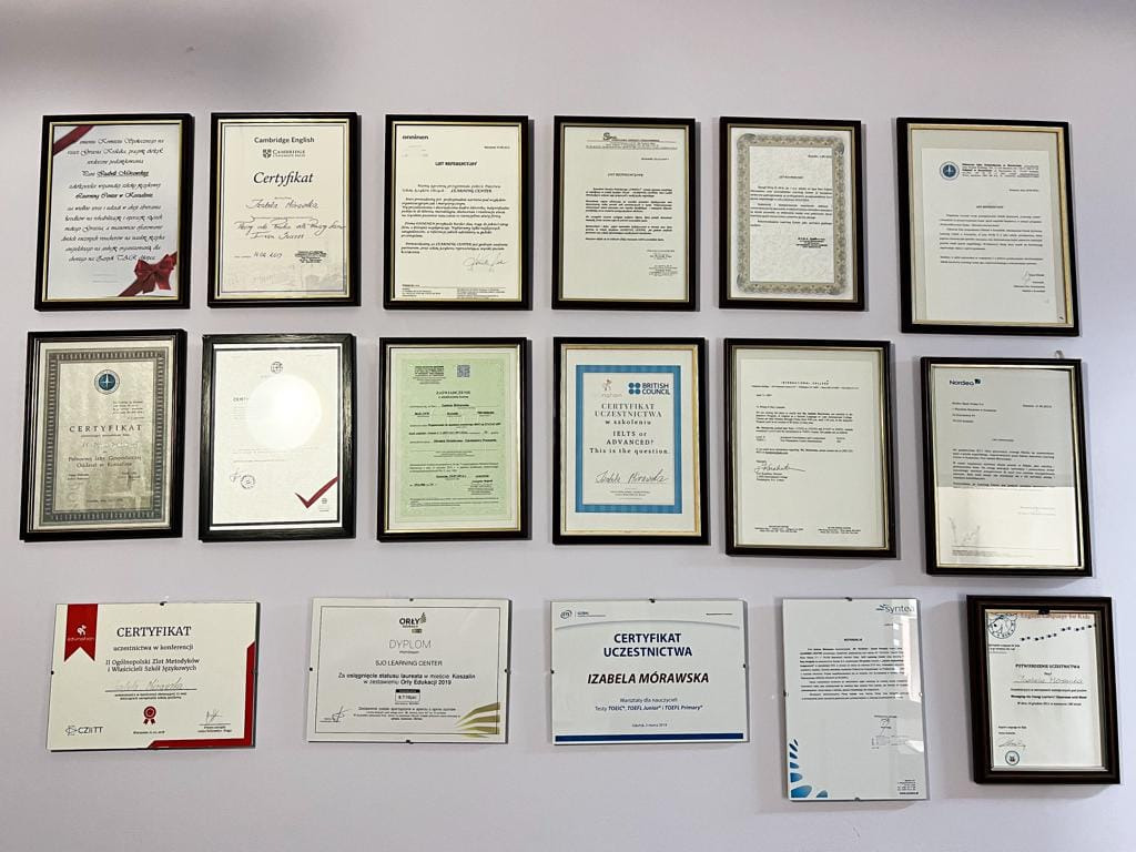 Ściana z certyfikatami i dyplomami w ramkach, potwierdzającymi kwalifikacje językowe, różnorodne wzory i formaty, oświetlenie dzienne, widoczne podpisy i pieczęcie.
