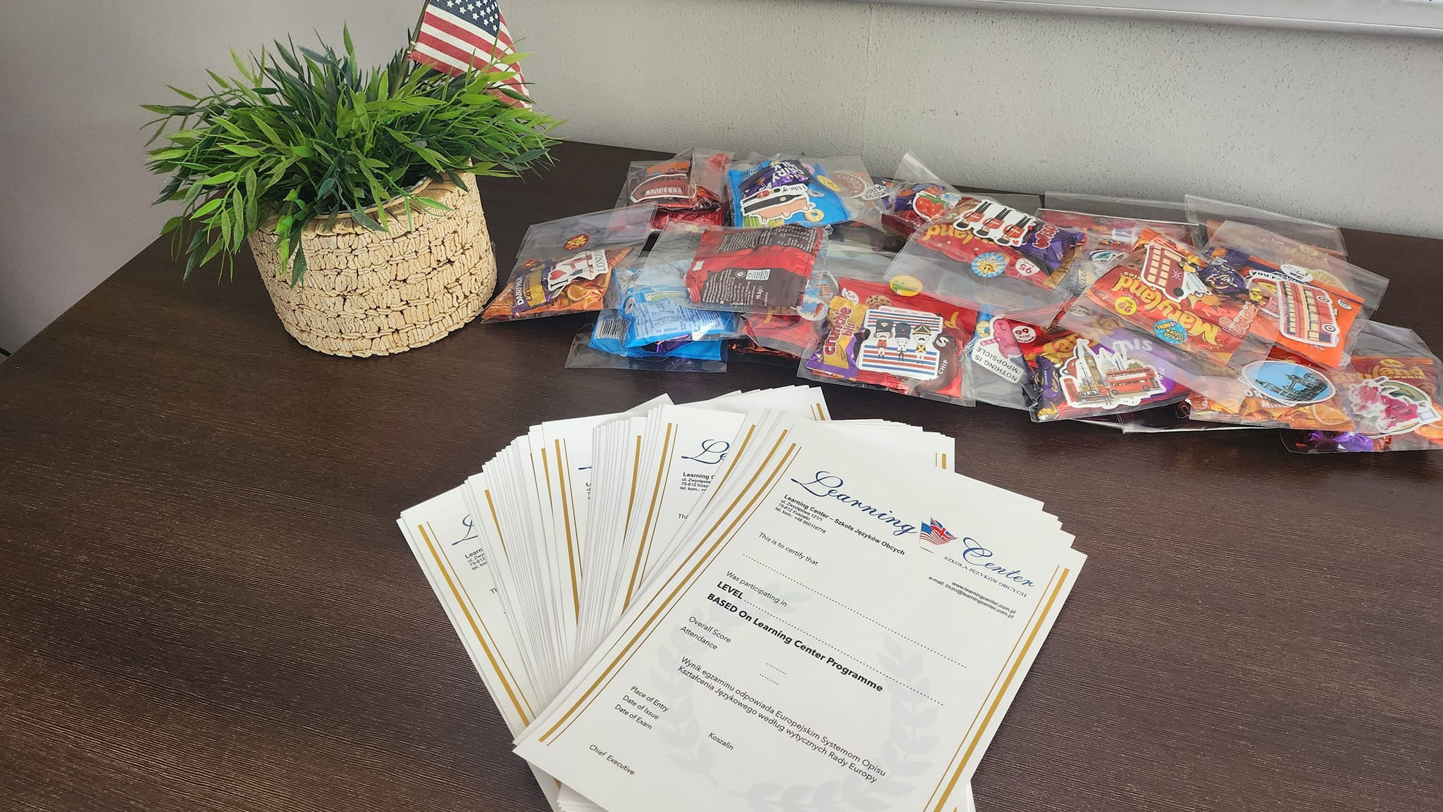Stół z certyfikatami ukończenia kursu językowego, słodyczami w torebkach z naklejkami i flagą USA. Nagroda za ukończenie kursu w szkole językowej.