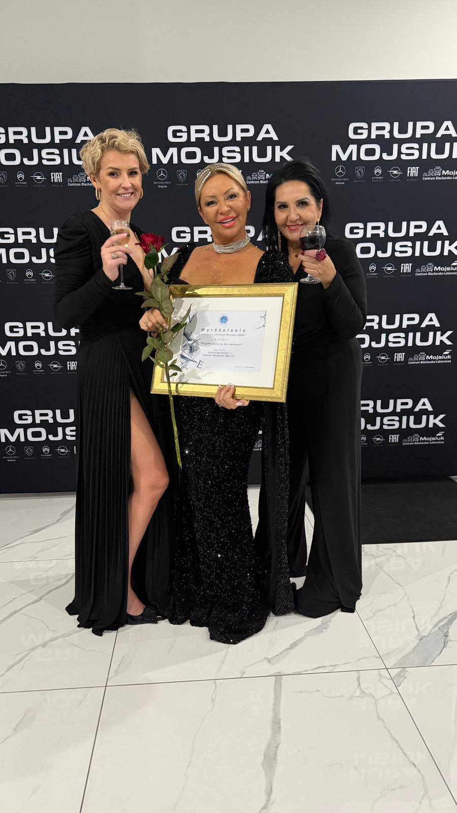 Trzy eleganckie kobiety w czarnych sukniach pozują z nagrodą 'Wyróżnienie' na tle logotypu 'Grupa Mojsiuk', trzymając kieliszki z winem i czerwoną różę.