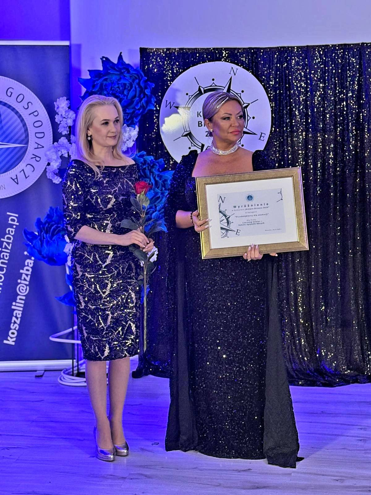 Uroczyste wręczenie wyróżnienia dla szkoły językowej w Koszalinie. Kobieta w cekinowej sukni trzyma oprawiony certyfikat na tle dekoracji.