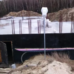 Wylany fundament domu z wystającymi prętami zbrojeniowymi, zabezpieczony czarną folią i ocieplony różowym styropianem, widoczny otwór wejściowy zabezpieczony płytą OSB oraz prowizoryczne okienko.