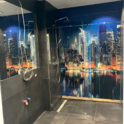Nowoczesna łazienka z czarnymi płytkami i kabiną prysznicową typu walk-in z fototapetą przedstawiającą panoramę miasta nocą, widoczne niedokończone podłączenia hydrauliczne.
