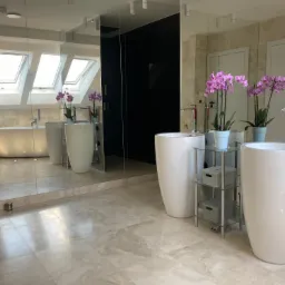 Elegancka łazienka z dwoma białymi umywalkami wolnostojącymi, marmurową podłogą, lustrem na całej ścianie, widoczną wanną i oknami dachowymi, ozdobiona fioletowymi storczykami.