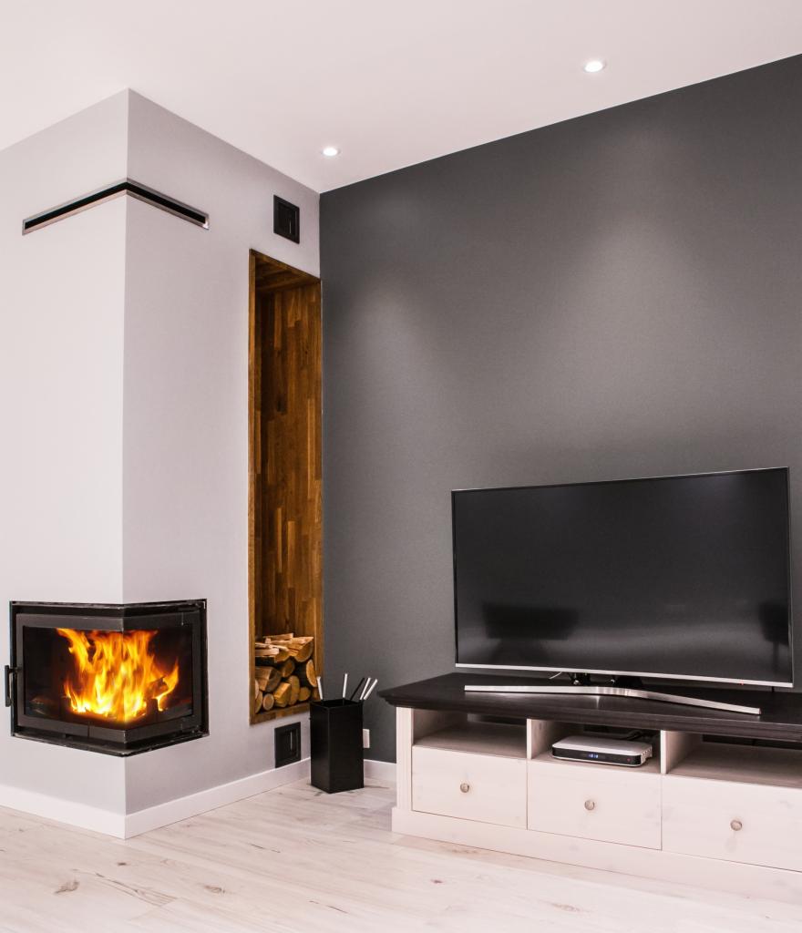 Minimalistyczny salon z narożnym kominkiem z palącym się ogniem, drewnianą wnęką na drewno i szarą ścianą z telewizorem.