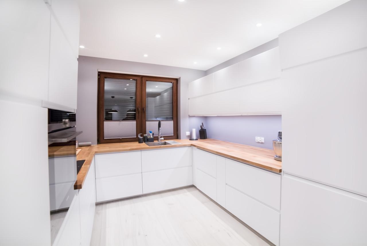 Nowoczesna kuchnia z białymi szafkami, drewnianym blatem i oknem w brązowej ramie z żaluzjami, oświetlona punktowymi lampami sufitowymi i listwą LED pod szafkami wiszącymi.