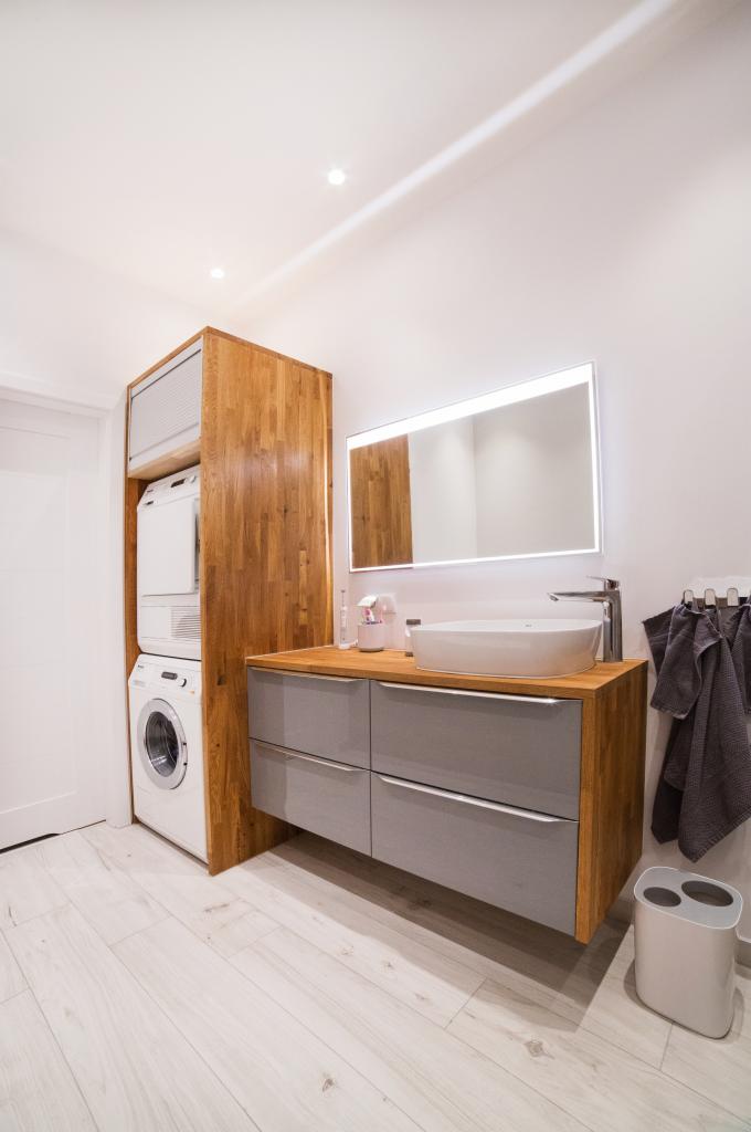 Nowoczesna łazienka z zabudowaną pralką i suszarką w drewnianej obudowie, szara szafka pod umywalką z drewnianym blatem i podświetlanym lustrem.