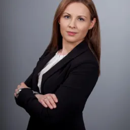 Portret kobiety w czarnym żakiecie i białej bluzce, z założonymi rękami na szarym tle, prezentujący profesjonalny wizerunek.