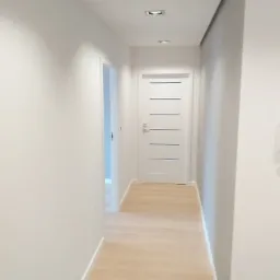 Wnętrze korytarza z jasną podłogą z paneli, białymi ścianami i drzwiami z poziomymi, ciemnymi wstawkami, widoczne dwa halogenowe punkty świetlne w suficie.