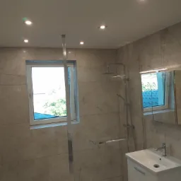 Wykończona łazienka z kabiną prysznicową typu walk-in, beżowe płytki imitujące marmur na ścianach, biała szafka z umywalką i lustrem, oświetlenie punktowe w suficie.