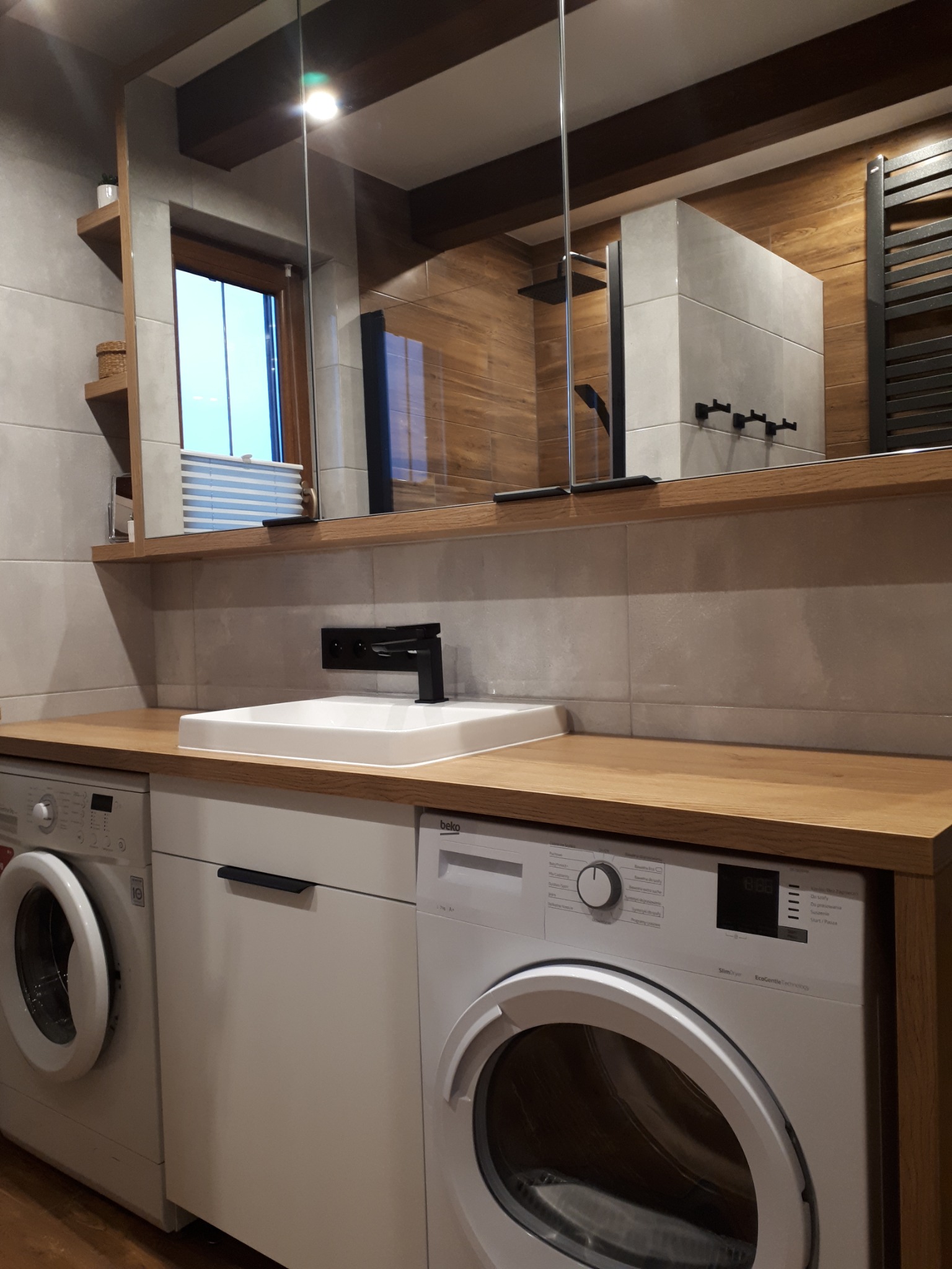 Nowoczesna łazienka z drewnianymi akcentami, dwie pralki pod blatem z umywalką i czarną armaturą, lustro z widokiem na kabinę prysznicową i grzejnik drabinkowy.