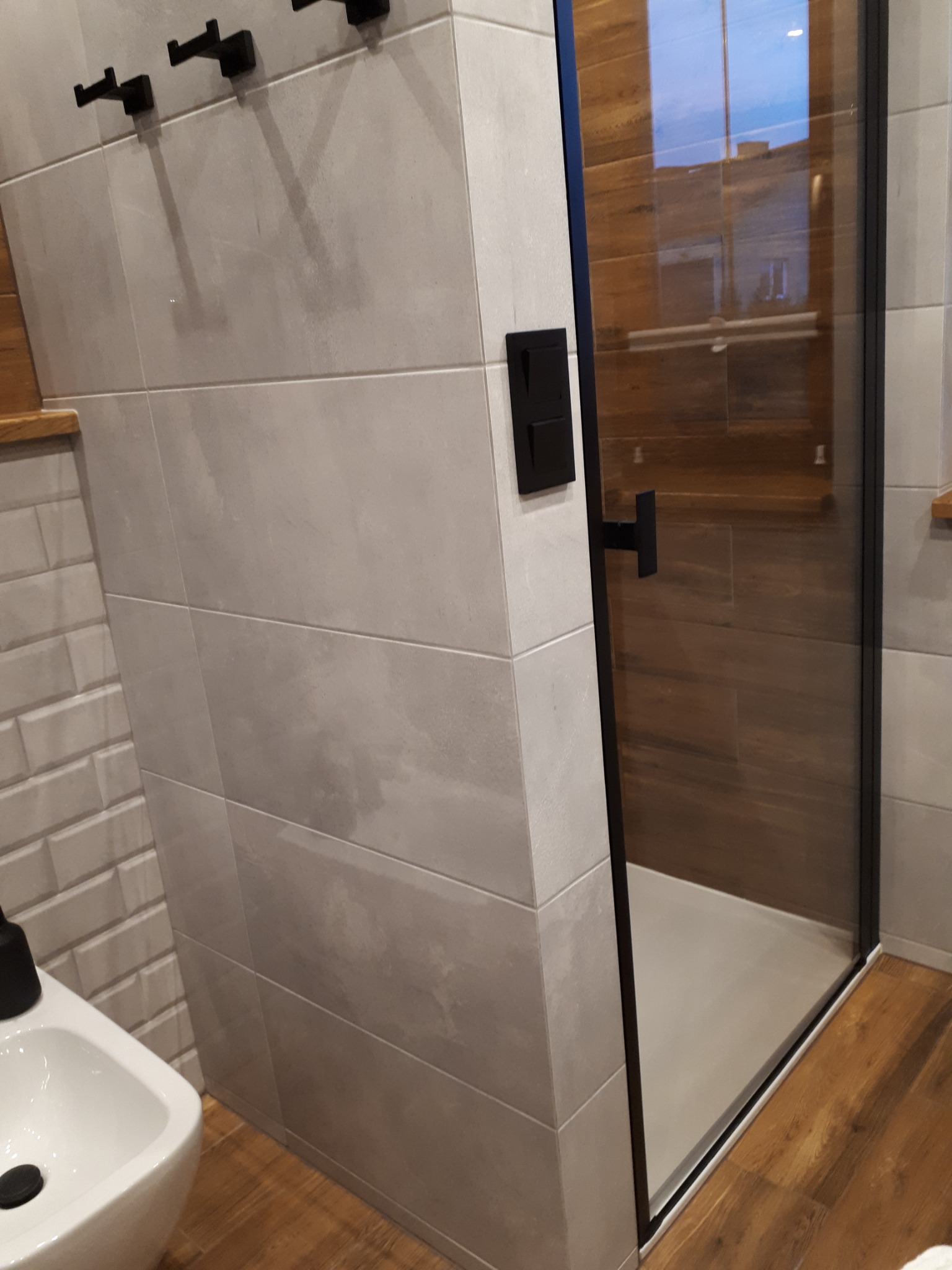 Nowoczesna łazienka z kabiną prysznicową typu walk-in, szare płytki imitujące beton na ścianach, drewnopodobne płytki wewnątrz kabiny i na podłodze, czarne akcenty w postaci wieszaków i armatury...
