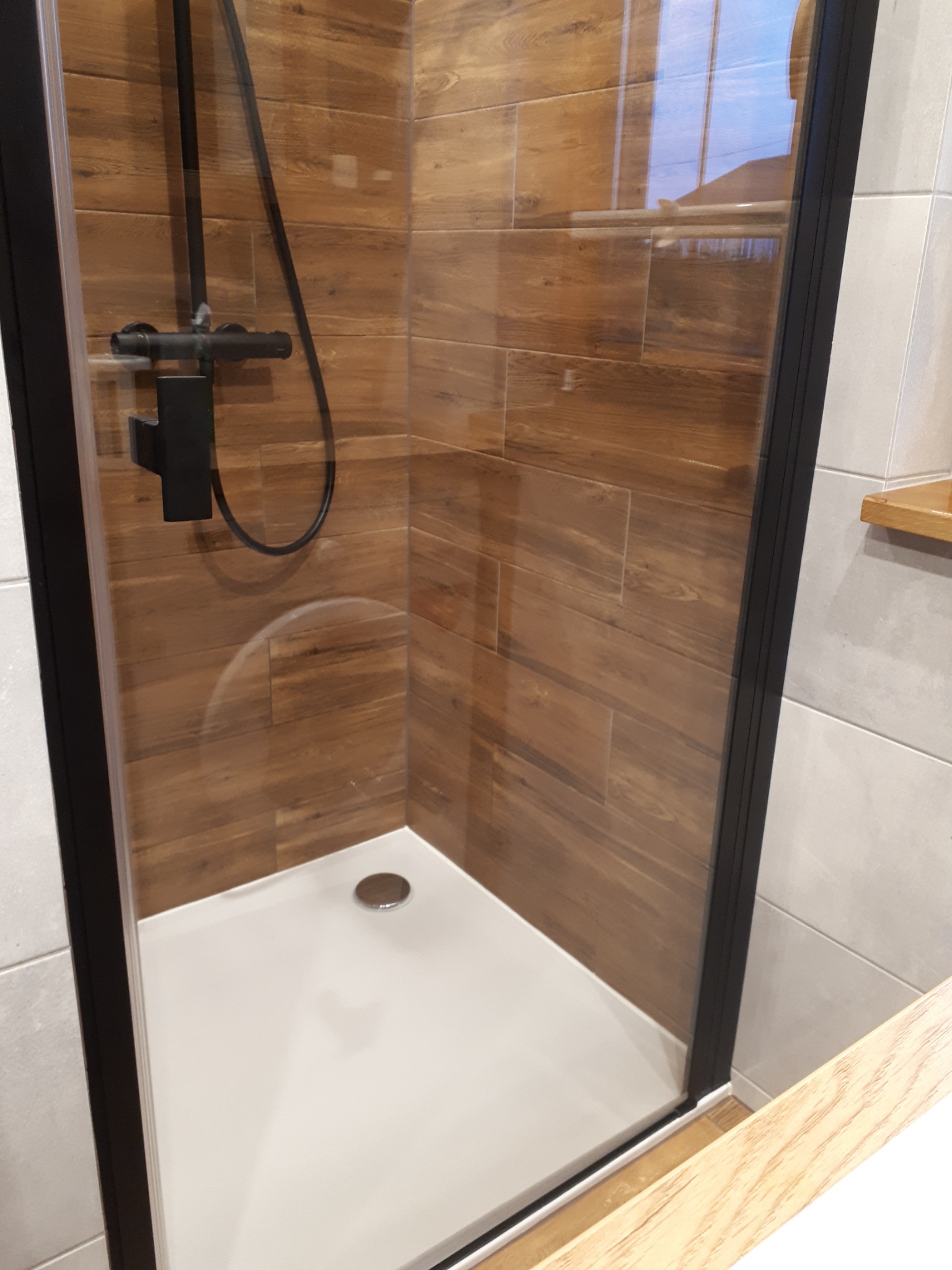 Kabina prysznicowa z czarną ramą i deszczownicą na tle drewnopodobnych płytek w łazience.