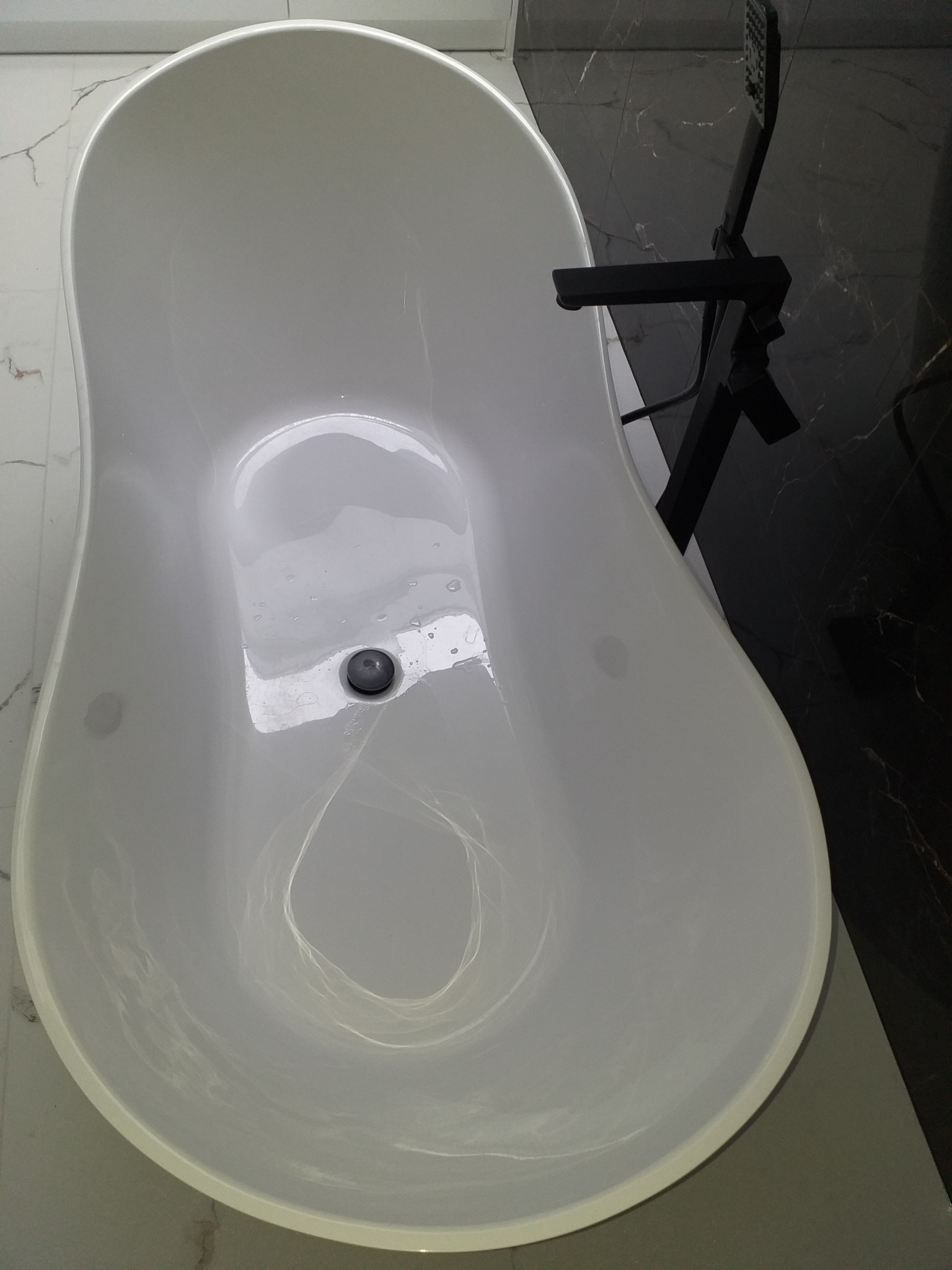 Biała, wolnostojąca wanna z czarną armaturą, widok z góry, marmurowe płytki w tle.