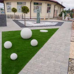Wzorcowa aranżacja terenu przed budynkiem z ekspozycją nawierzchni z szarej kostki brukowej, trawnika z białymi kulami dekoracyjnymi i elementów z kamienia ozdobnego.