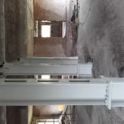 Białe, pomalowane metalowe słupy konstrukcyjne w surowym wnętrzu budynku z ceglanymi ścianami i betonową podłogą, widoczne ślady niedokończonych prac.