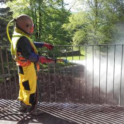 Firma Usługowa REMAX Leszek Świątecki - Pracownik w kombinezonie ochronnym i hełmie piaskuje metalową balustradę na balkonie, strumień ścierniwa tworzy obłok pyłu, w tle widoczne drzewa.