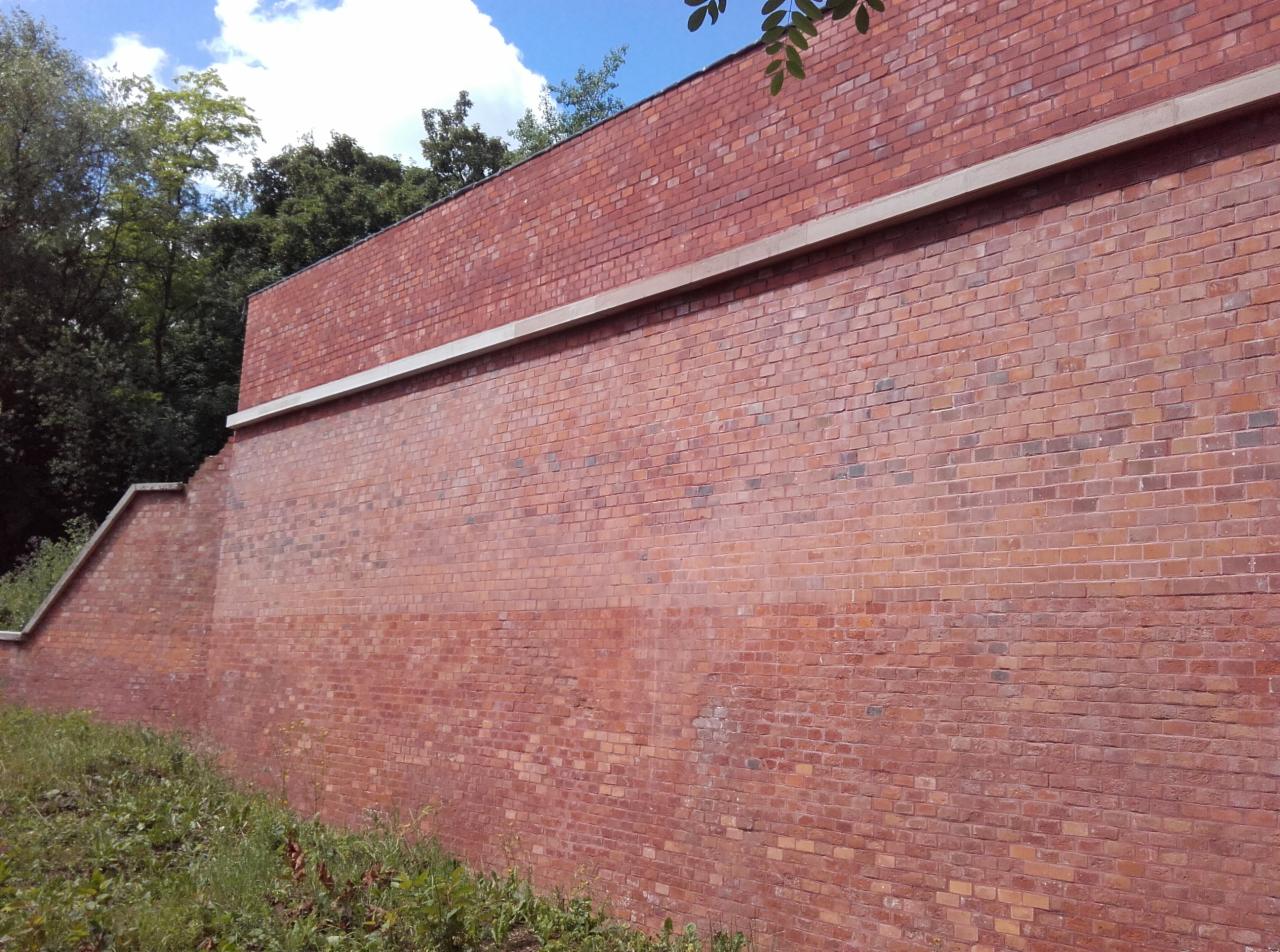 Ceglany mur oporowy z betonowym zwieńczeniem, częściowo porośnięty roślinnością, widoczny z dołu pod kątem.