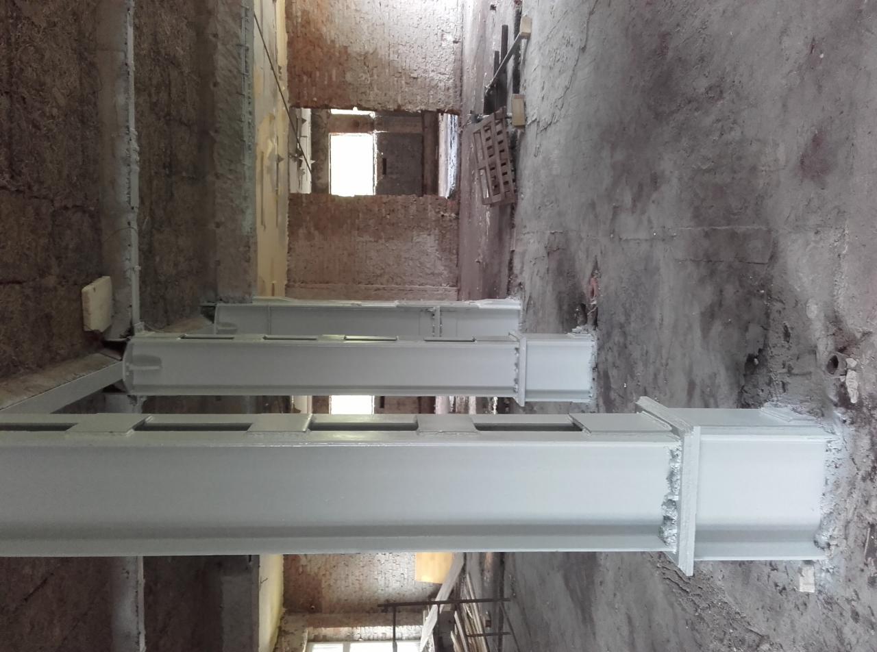 Białe, pomalowane metalowe słupy konstrukcyjne w surowym wnętrzu budynku z ceglanymi ścianami i betonową podłogą, widoczne ślady niedokończonych prac.