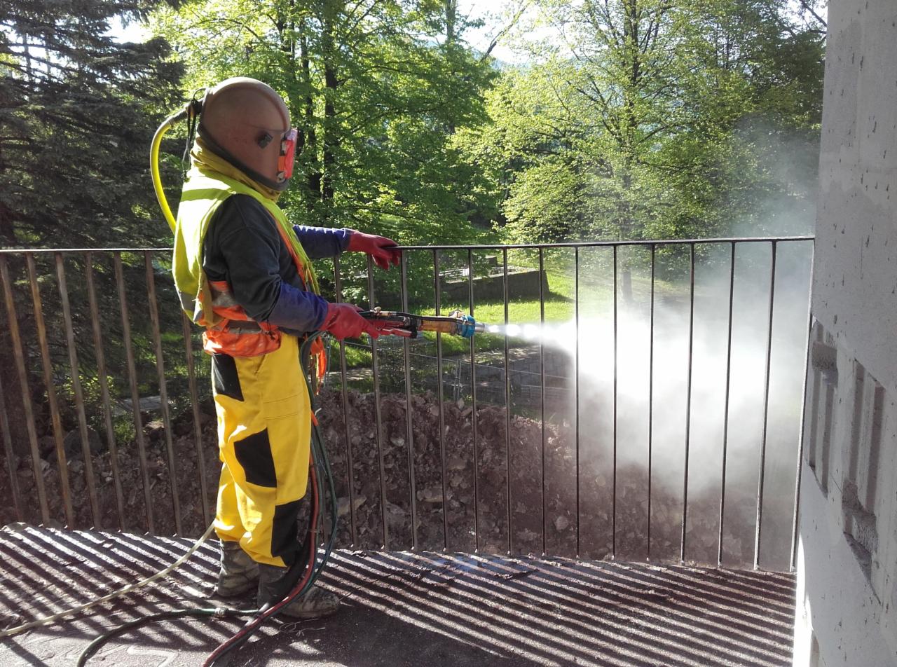 Pracownik w kombinezonie ochronnym i hełmie piaskuje metalową balustradę na balkonie, strumień ścierniwa tworzy obłok pyłu, w tle widoczne drzewa.
