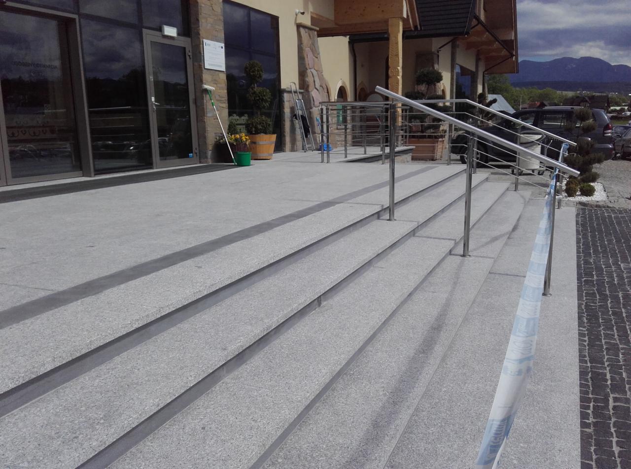 Schody wejściowe do budynku wyłożone szarym, ziarnistym kamieniem z metalową barierką, widok na drzwi wejściowe, rośliny w donicach i fragment gór w tle.