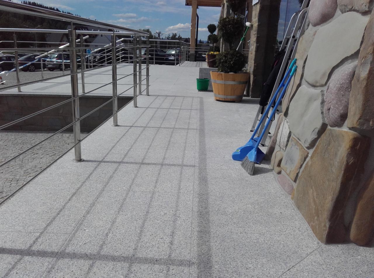 Szara, granitowa posadzka tarasu z metalową balustradą, drewnianą donicą z roślinami, oparta o kamienną ścianę. Dwie niebieskie zmiotki oparte o ścianę.