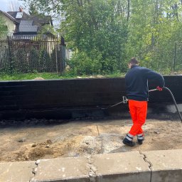 Budowy-Remonty Robert Niewiadomski - Pracownik w pomarańczowych spodniach zabezpiecza fundamenty budynku smolistą substancją za pomocą lancy natryskowej, widoczne świeżo zaizolowane czarne ściany fundamentowe z cegły.