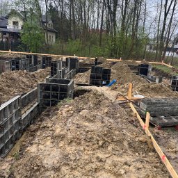 Budowy-Remonty Robert Niewiadomski - Budowa fundamentów pod dom jednorodzinny: szalunki zalane błotem, wystające pręty zbrojeniowe, w tle drzewa i fragment zabudowy mieszkalnej.