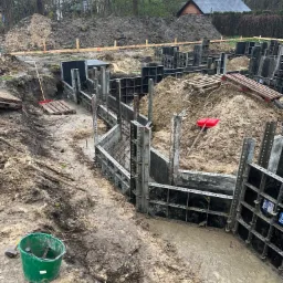 Wykop pod fundamenty domu z użyciem szalunków systemowych, widoczny zielony kubeł z narzędziami i pryzma ziemi w tle.