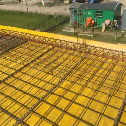 Zbrojenie stropu na żółtym szalunku, widoczne pionowe pręty zbrojeniowe, w tle barak budowlany i toaleta przenośna.