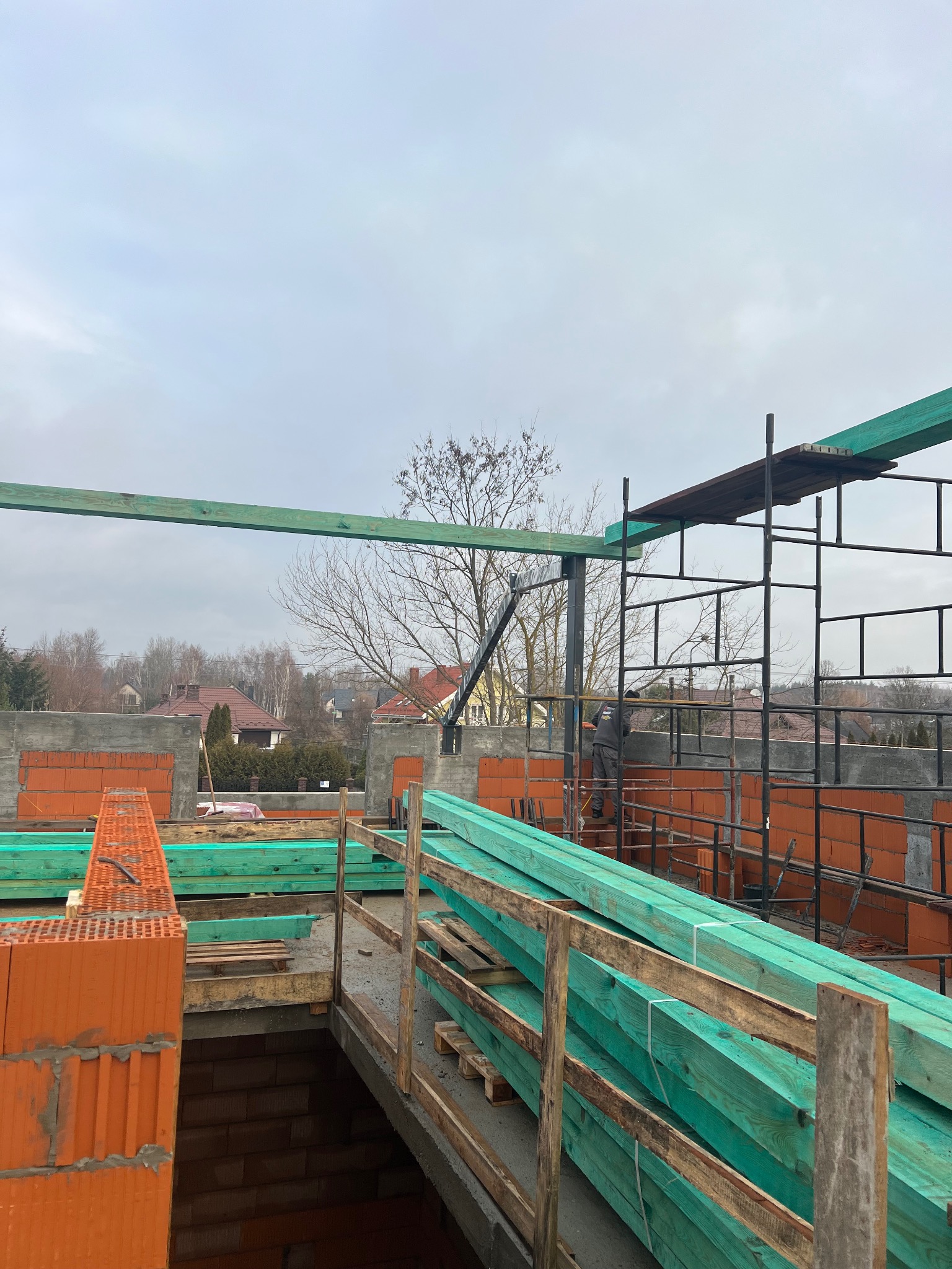 Wznoszenie ścian z czerwonej cegły na budowie domu jednorodzinnego, widoczne rusztowania i przygotowane drewniane elementy konstrukcyjne w kolorze zielonym, pochmurne niebo w tle.