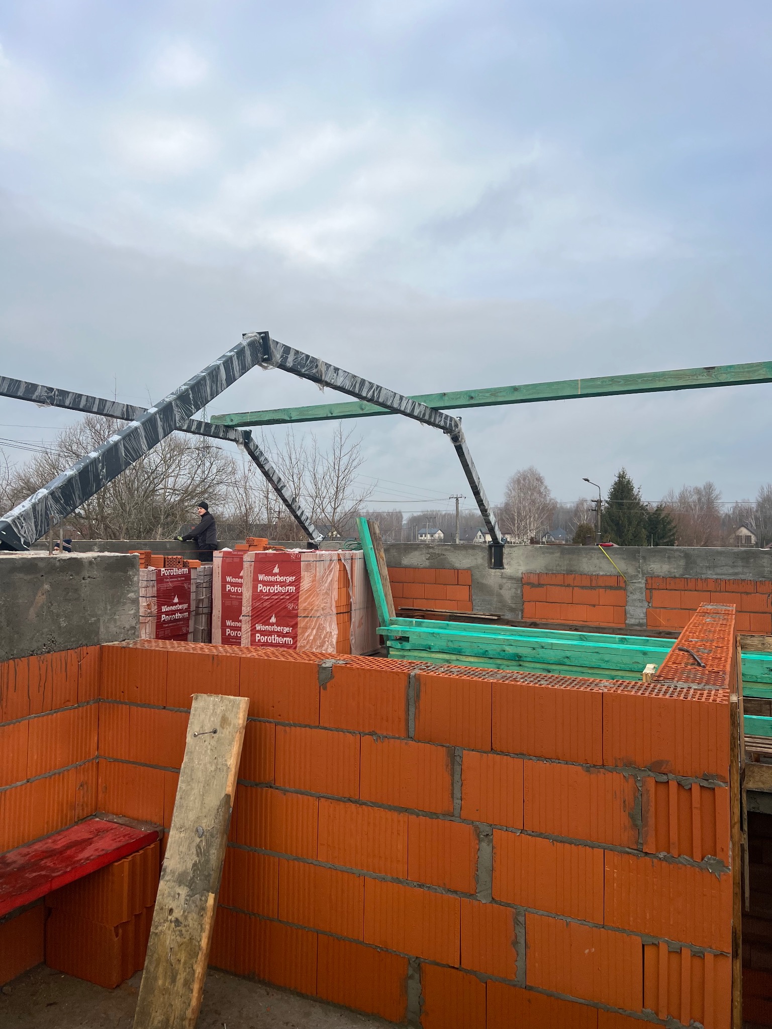 Wznoszenie ścian parteru z czerwonej ceramiki poryzowanej, widoczne palety z zapakowanymi bloczkami, konstrukcja dachu w trakcie montażu, pracownik w oddali.