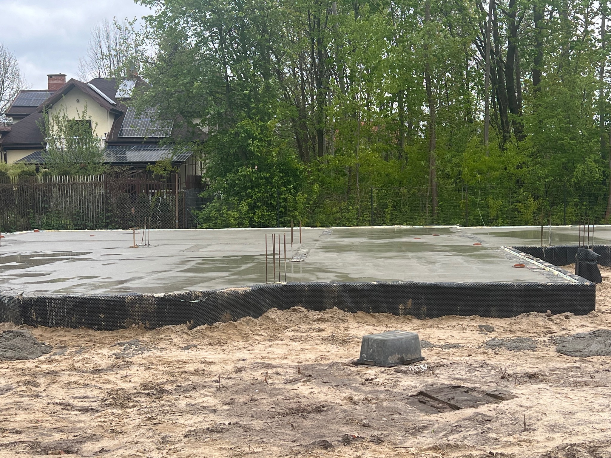 Wylana, mokra płyta fundamentowa z wystającymi prętami zbrojeniowymi, zabezpieczona czarną folią izolacyjną na piaszczystym gruncie; w tle widoczny dom z panelami słonecznymi na dachu i drzewa.