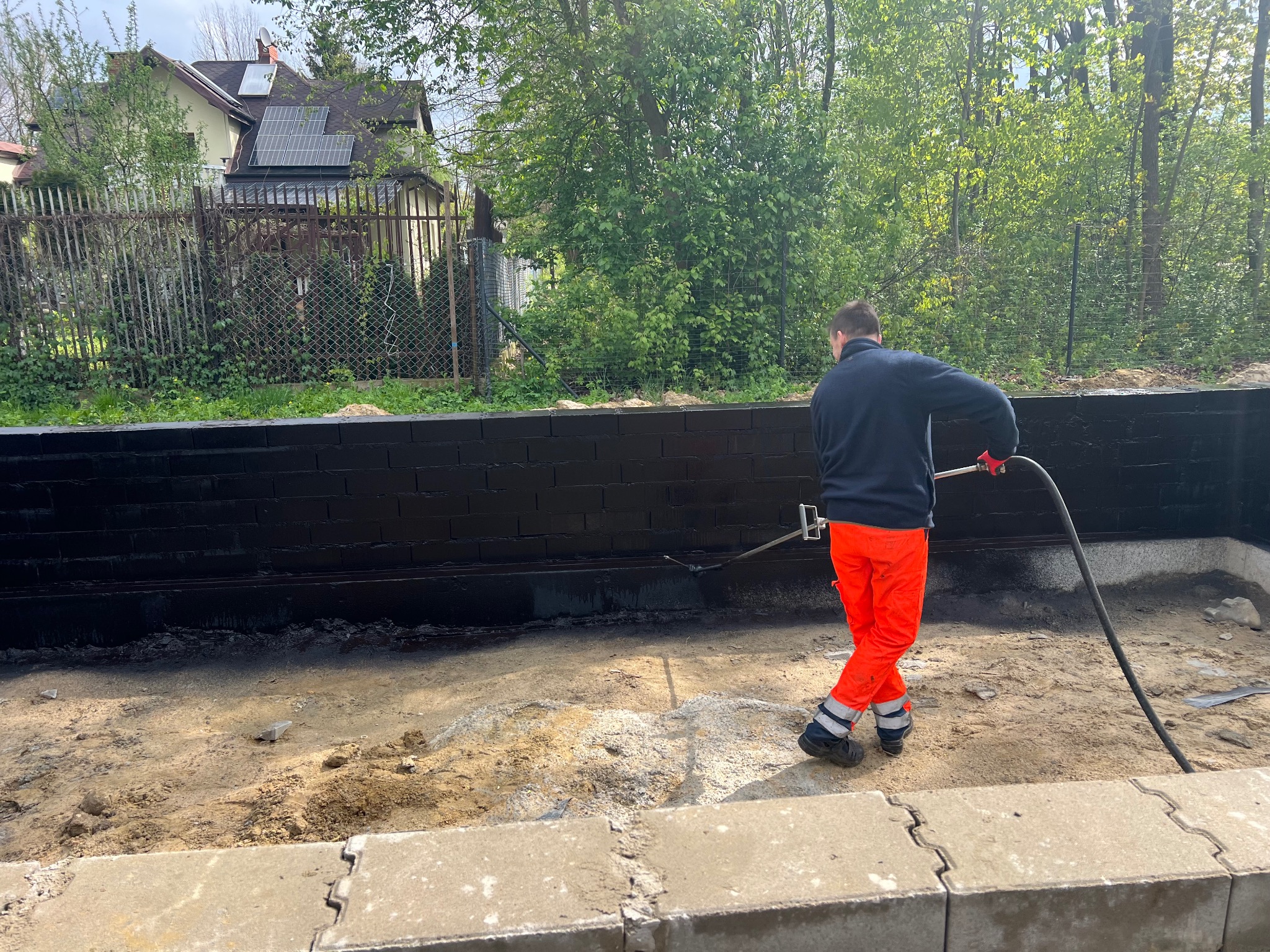 Pracownik w pomarańczowych spodniach zabezpiecza fundamenty budynku smolistą substancją za pomocą lancy natryskowej, widoczne świeżo zaizolowane czarne ściany fundamentowe z cegły.