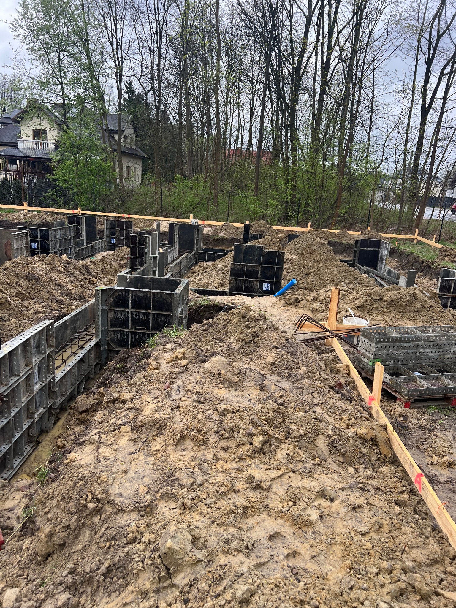Budowa fundamentów pod dom jednorodzinny: szalunki zalane błotem, wystające pręty zbrojeniowe, w tle drzewa i fragment zabudowy mieszkalnej.