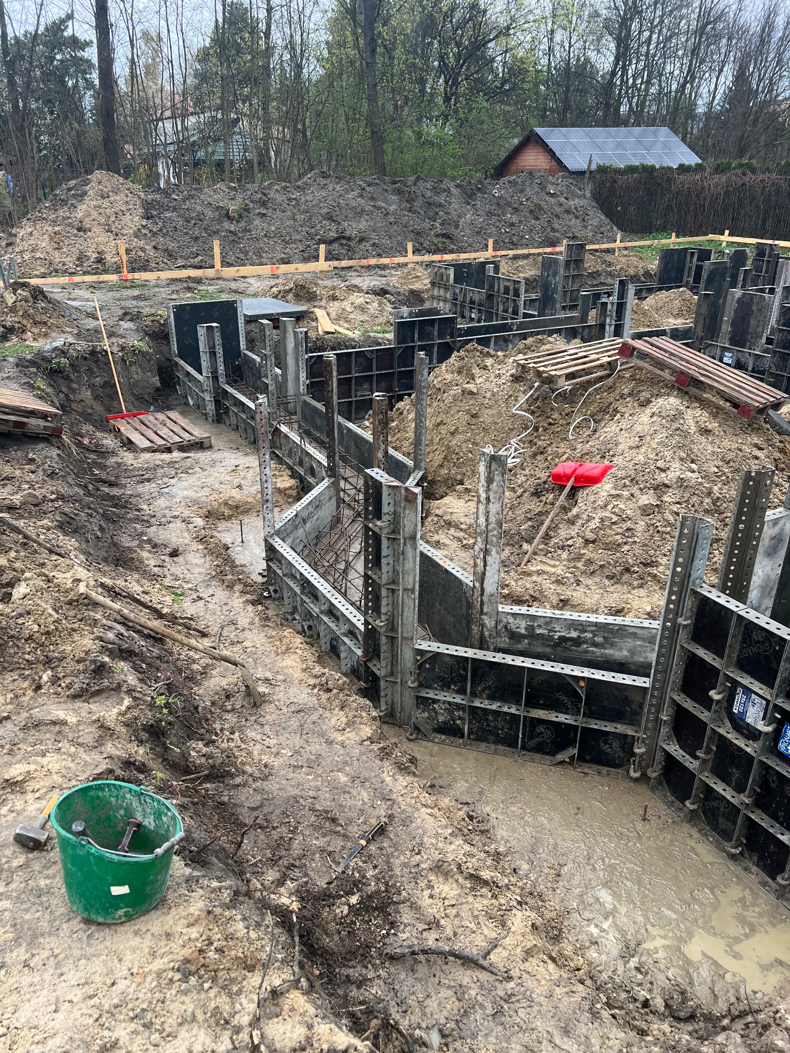 Wykop pod fundamenty domu z użyciem szalunków systemowych, widoczny zielony kubeł z narzędziami i pryzma ziemi w tle.