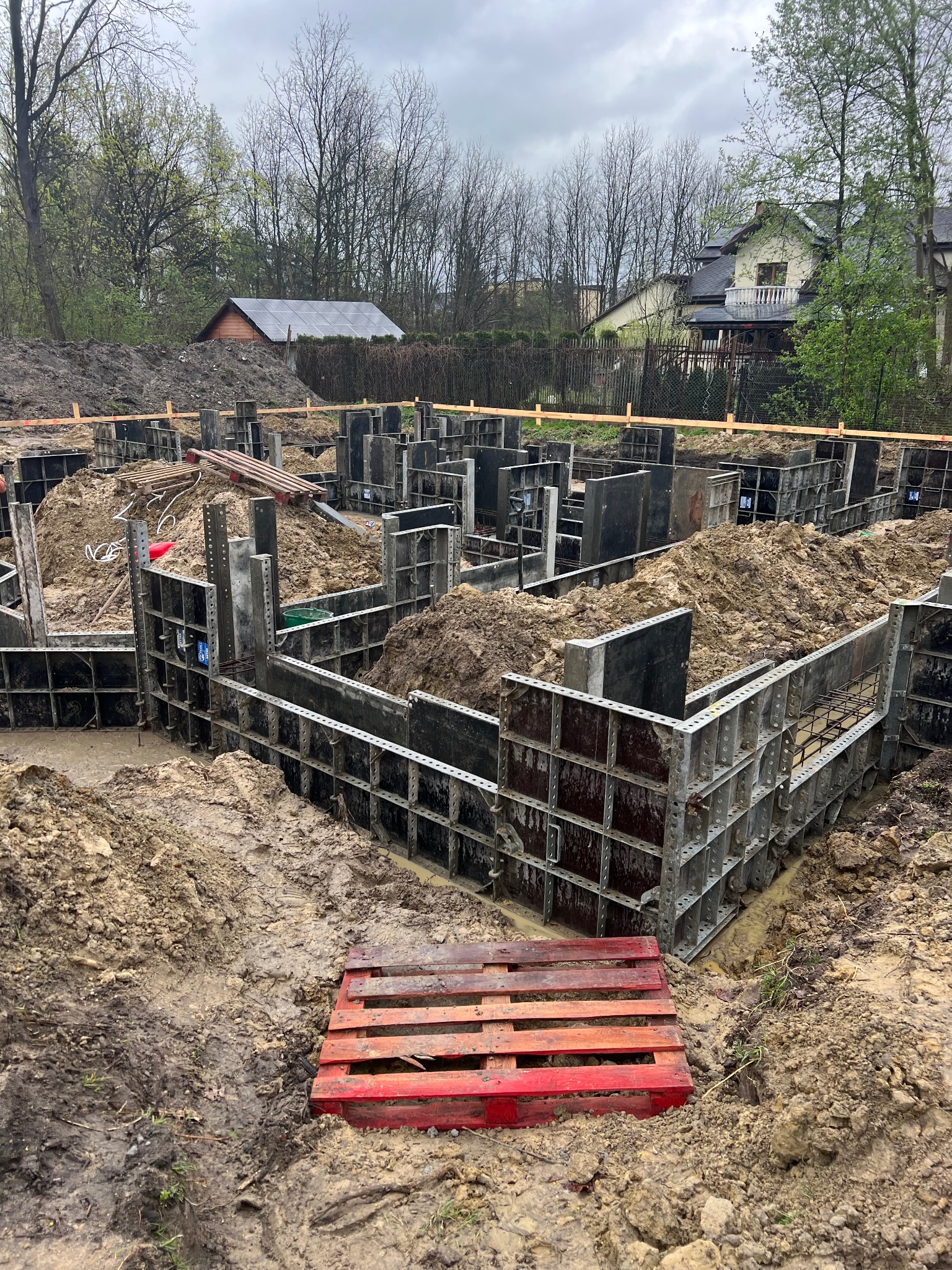 Przygotowanie fundamentów pod budowę domu: metalowe szalunki zalane ziemią, czerwona paleta na pierwszym planie, w tle widoczny dom i drzewa.