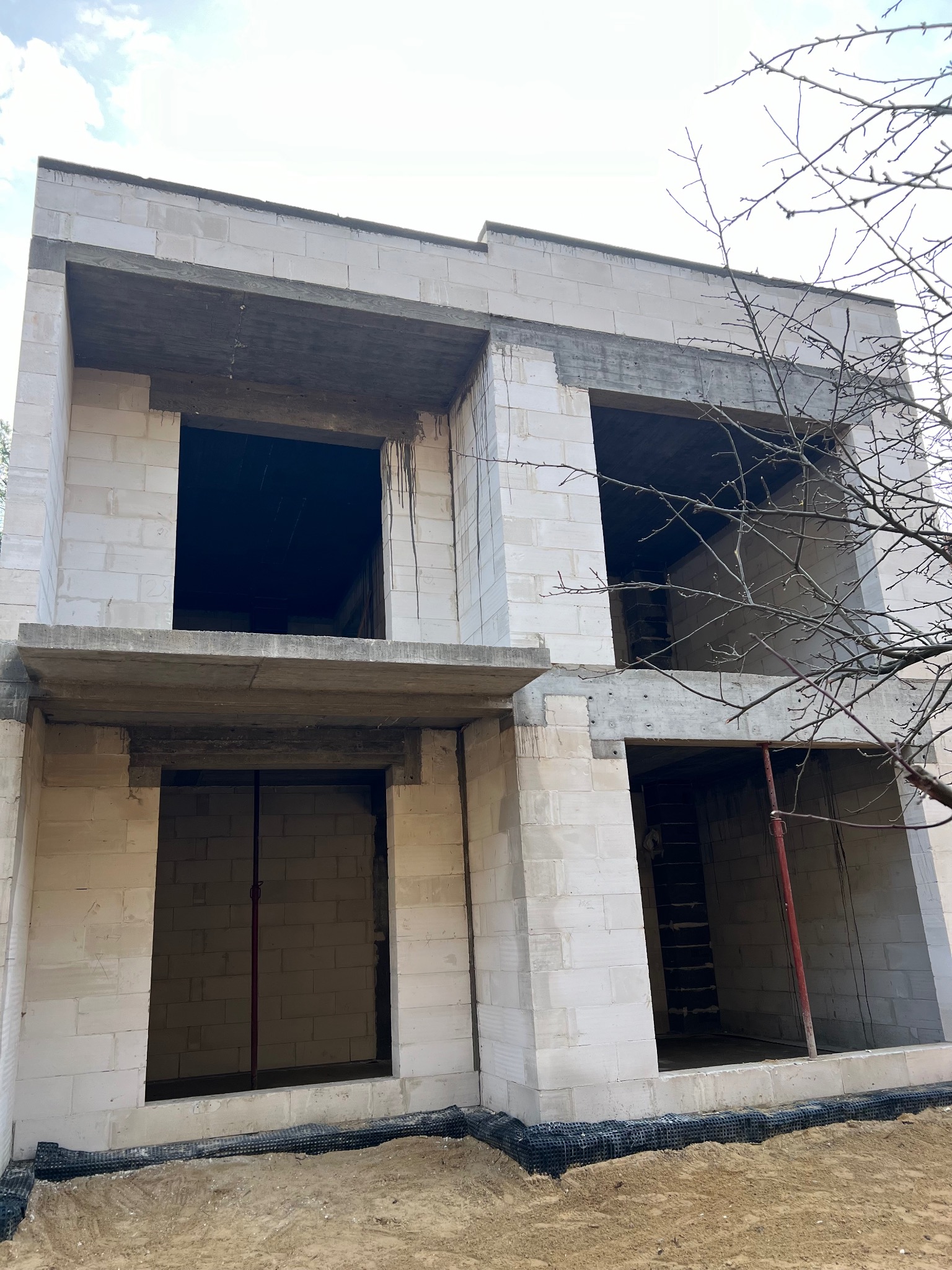 Surowy stan dwukondygnacyjnego domu z białych bloczków, widoczne otwory okienne i drzwiowe, teren wokół budynku przygotowany pod dalsze prace.