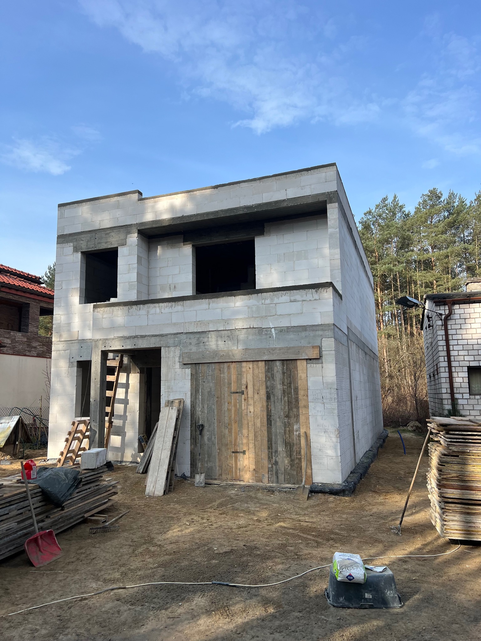 Dwukondygnacyjny budynek w stanie surowym otwartym z betonu komórkowego, z drewnianymi deskami zabezpieczającymi otwór garażowy, widoczny na tle błękitnego nieba i drzew