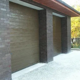 Dwa garaże z brązowymi, segmentowymi bramami, wkomponowane w ceglane filary i ściany, z widocznymi śladami wilgoci na bramach, otoczone kostką brukową i zielenią.