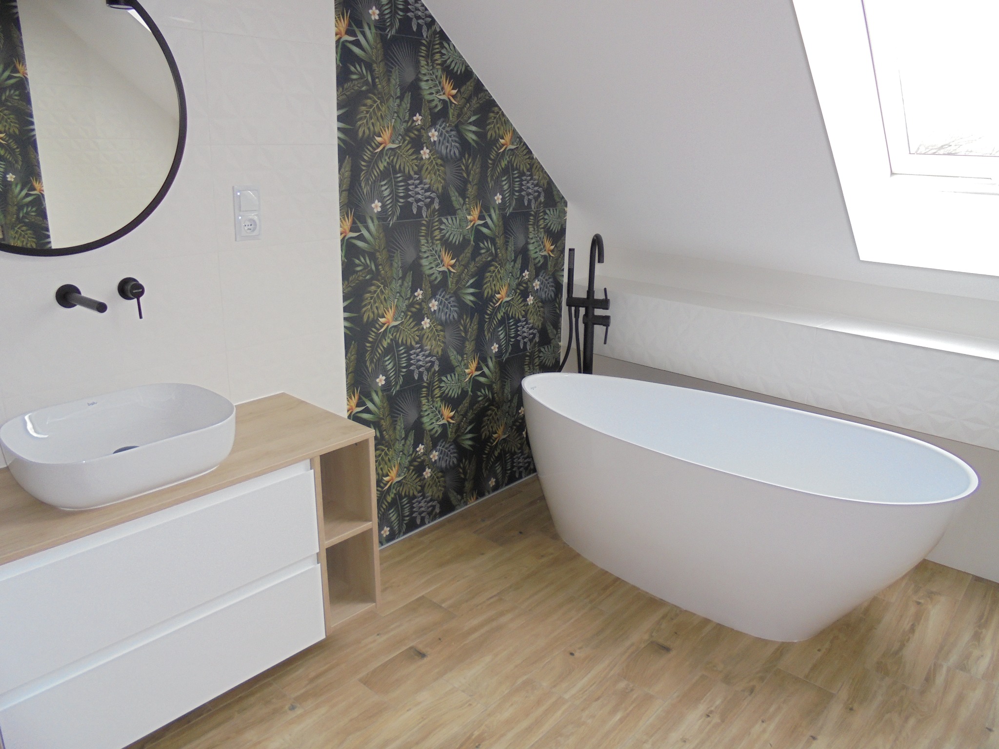 Nowoczesna łazienka na poddaszu z wolnostojącą białą wanną, umywalką nablatową na szafce z drewnianym blatem, okrągłym lustrem i ścianą w tropikalne wzory.