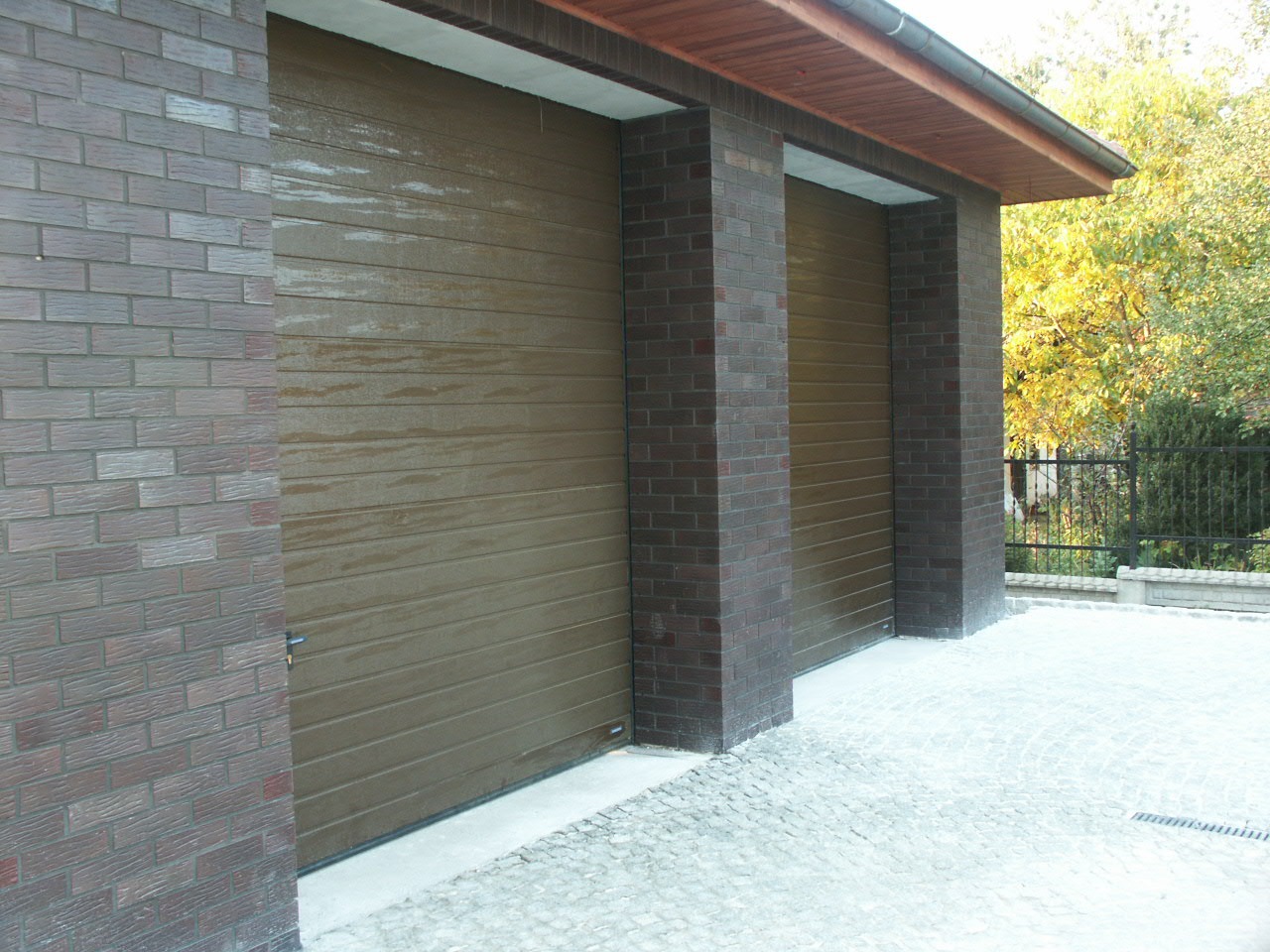 Dwa garaże z brązowymi, segmentowymi bramami, wkomponowane w ceglane filary i ściany, z widocznymi śladami wilgoci na bramach, otoczone kostką brukową i zielenią.