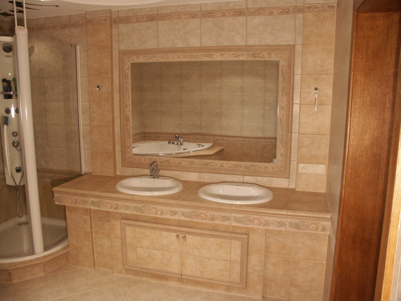 Jasna łazienka z beżowymi płytkami, kabiną prysznicową z hydromasażem, dwoma umywalkami wpuszczonymi w blat z dekoracyjnym frezem i dużym lustrem w ozdobnej ramie. Widoczna wanna w lustrzanym odbiciu.