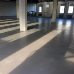 Szara, gładka posadzka w dużym pomieszczeniu, oświetlona promieniami słonecznymi wpadającymi przez okna, tworzącymi wyraźne pasy światła i cienia na powierzchni, z widocznymi białymi kolumnami...