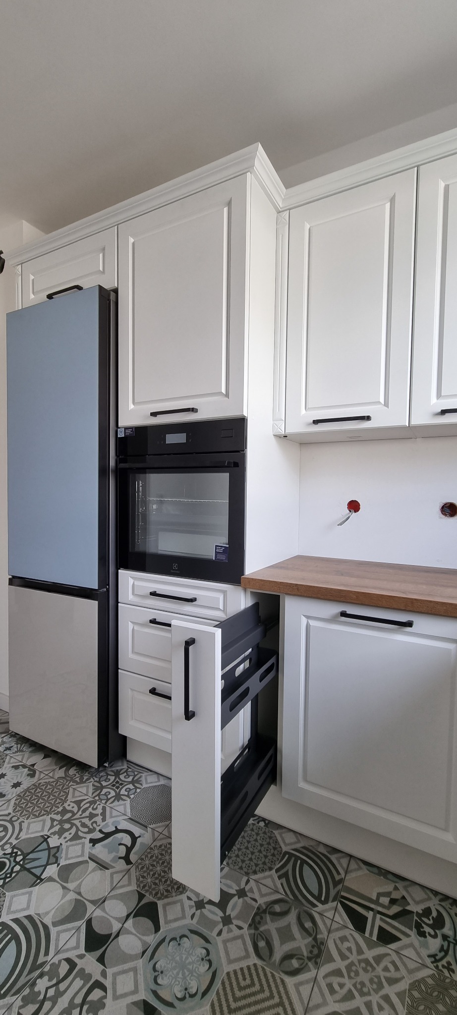 Biała zabudowa kuchenna z szufladą cargo, niebieska lodówka i wzorzysta podłoga z płytek.