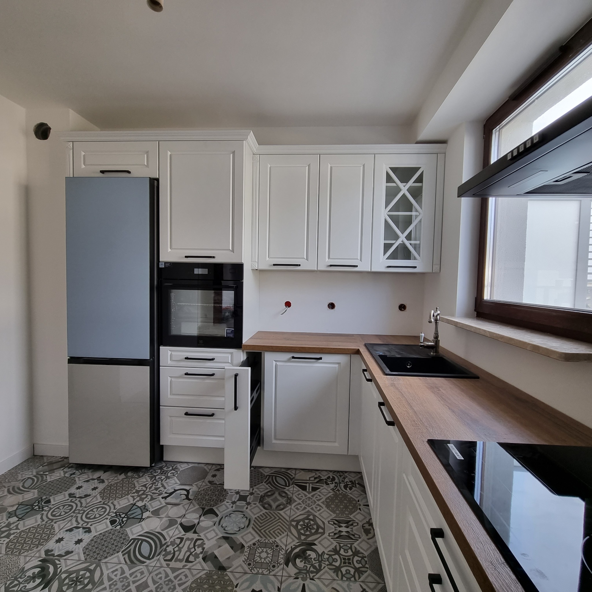 Jasna kuchnia z białymi szafkami z frezowanymi frontami, drewnianym blatem i wzorzystą podłogą, widoczny wysuwany kosz cargo i otwory w ścianie po instalacji elektrycznej.