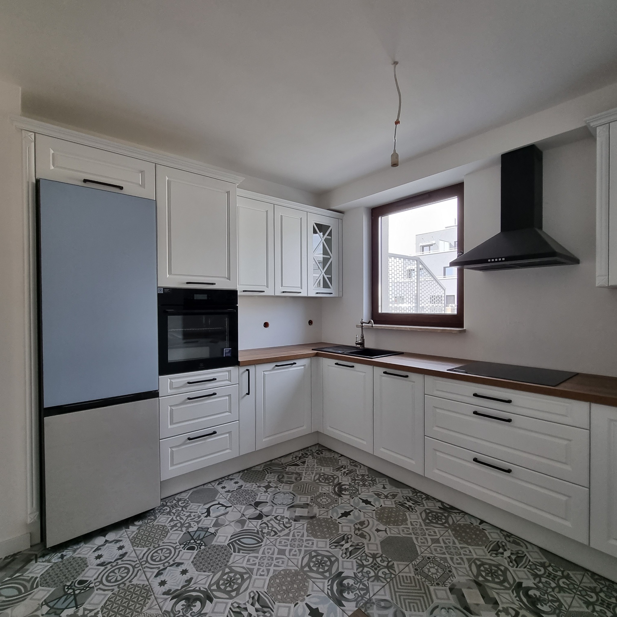 Nowoczesna kuchnia w bieli z szarym lodówką i wzorzystą podłogą, czarny okap nad płytą indukcyjną, drewniany blat i czarny zlew.