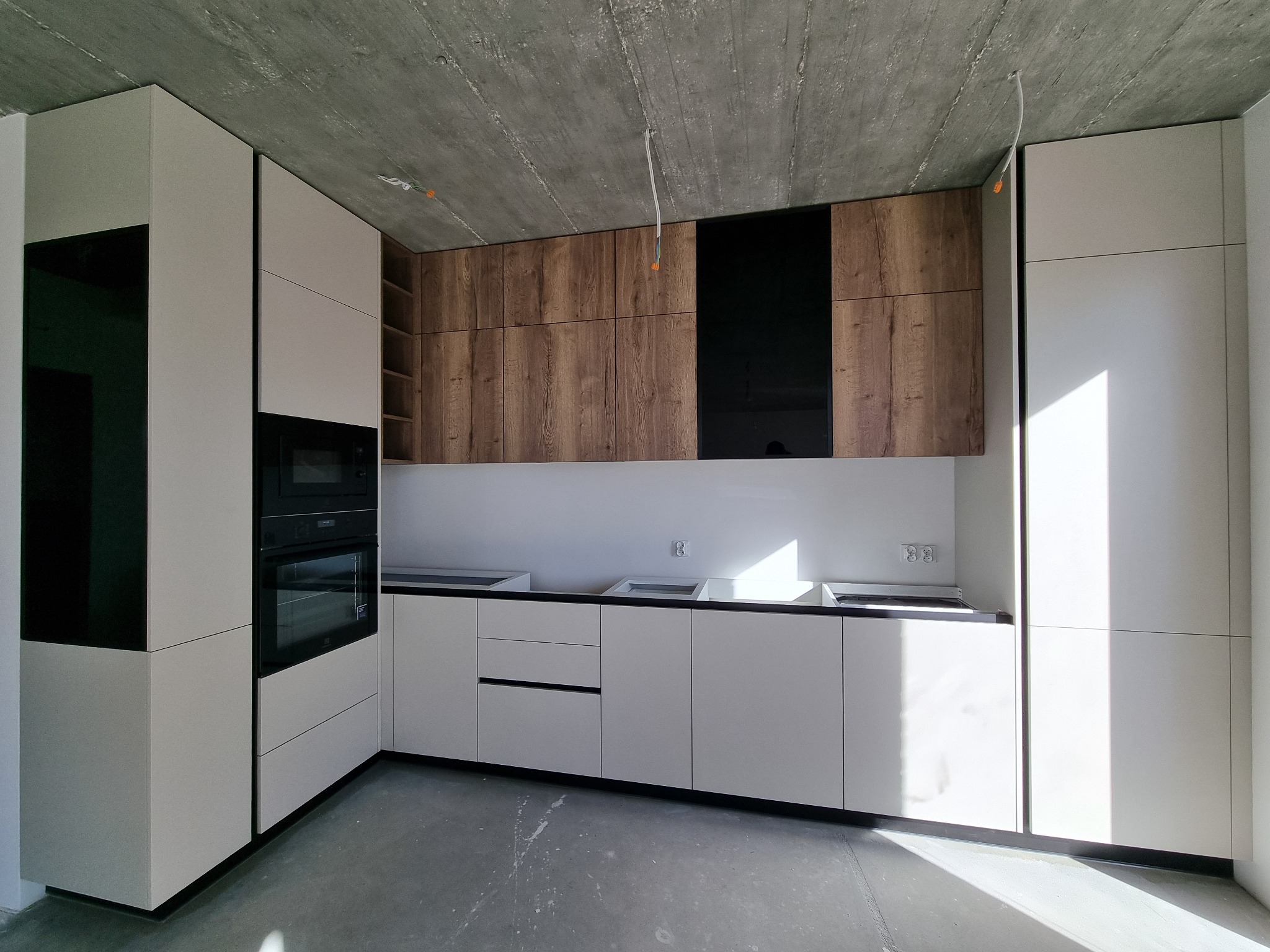 Nowoczesna kuchnia z białymi szafkami dolnymi i drewnianymi górnymi z czarnym akcentem, surowy betonowy sufit z widocznymi przewodami, w trakcie montażu.