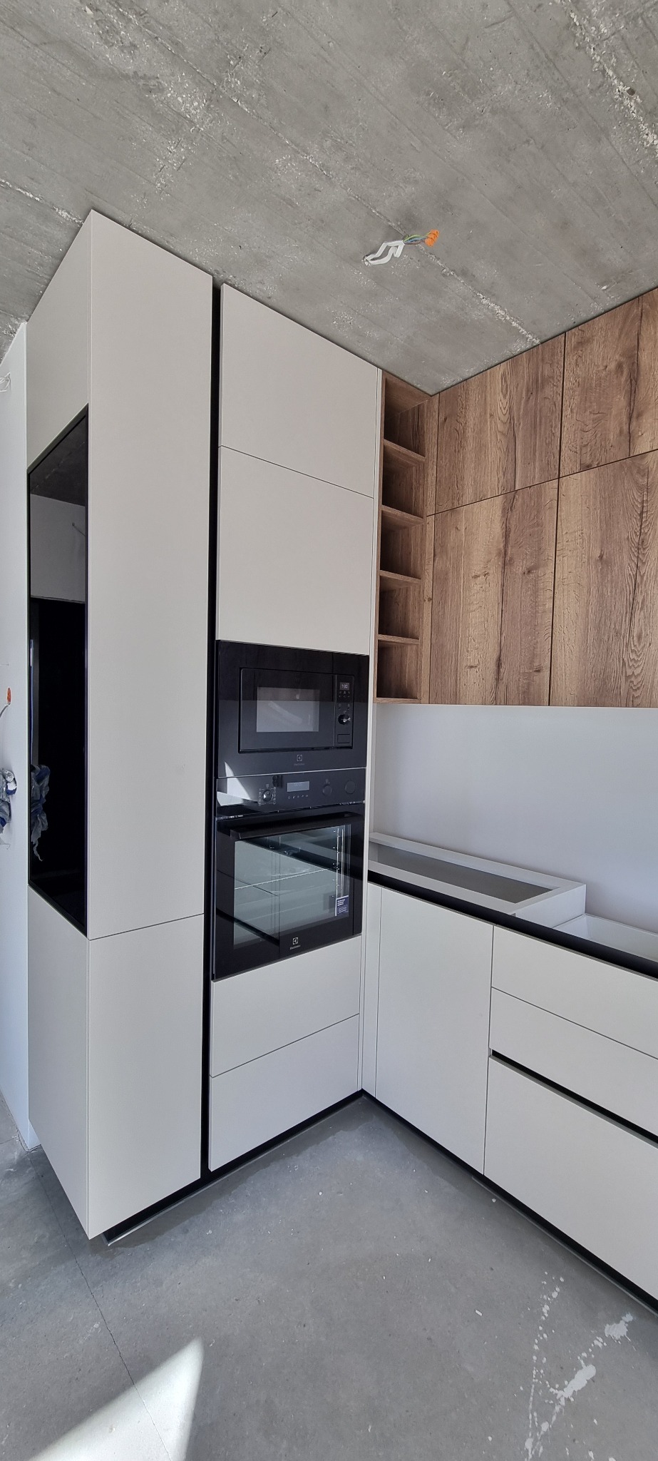 Nowoczesna kuchnia z białymi frontami i drewnianymi akcentami, zabudowany piekarnik i mikrofalówka Electrolux, surowy betonowy sufit z widocznymi przewodami.