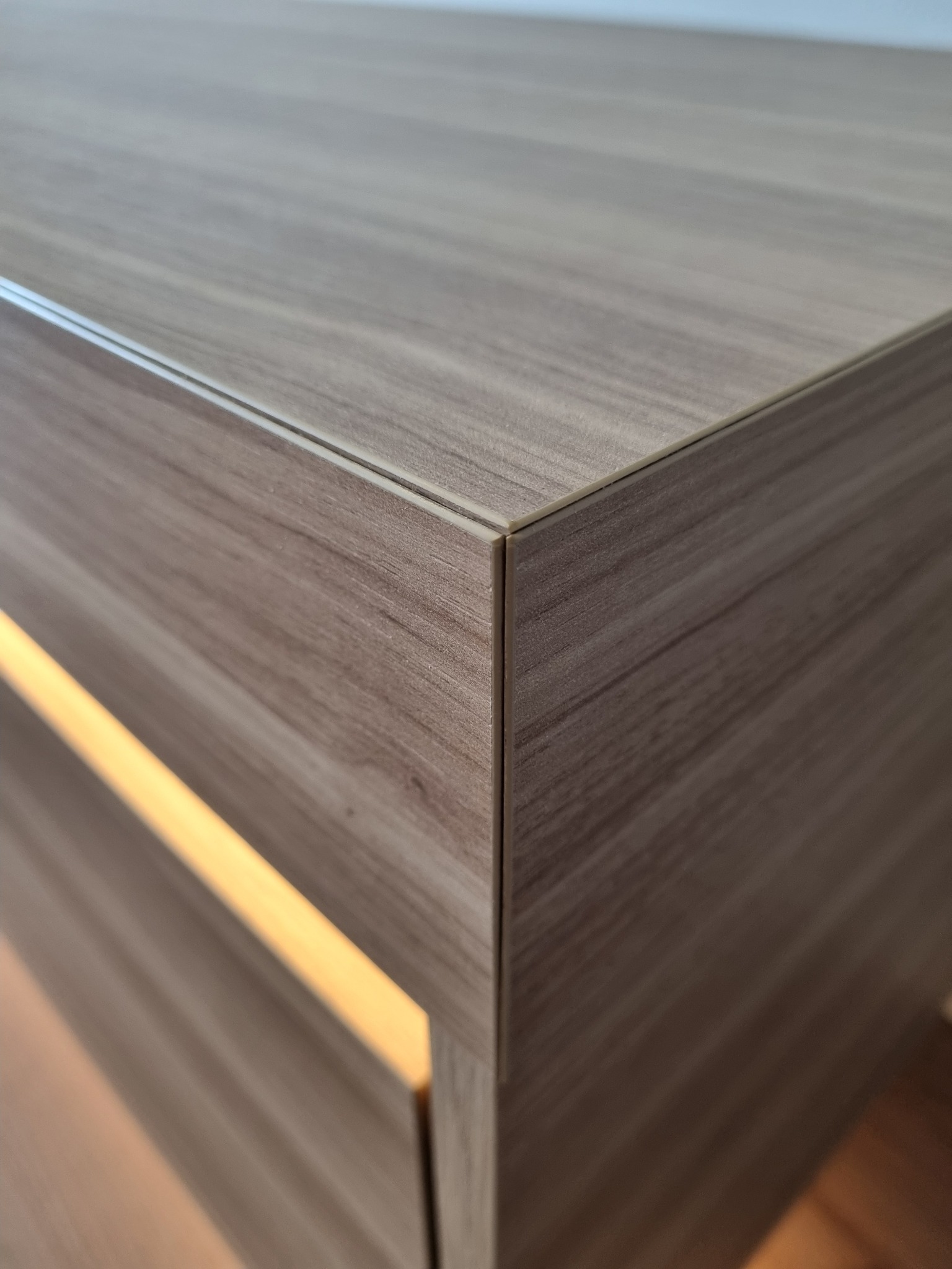 Fragment szafki z płyty laminowanej imitującej drewno, z subtelnym podświetleniem LED w szczelinie, ukazujący precyzyjne łączenie krawędzi.