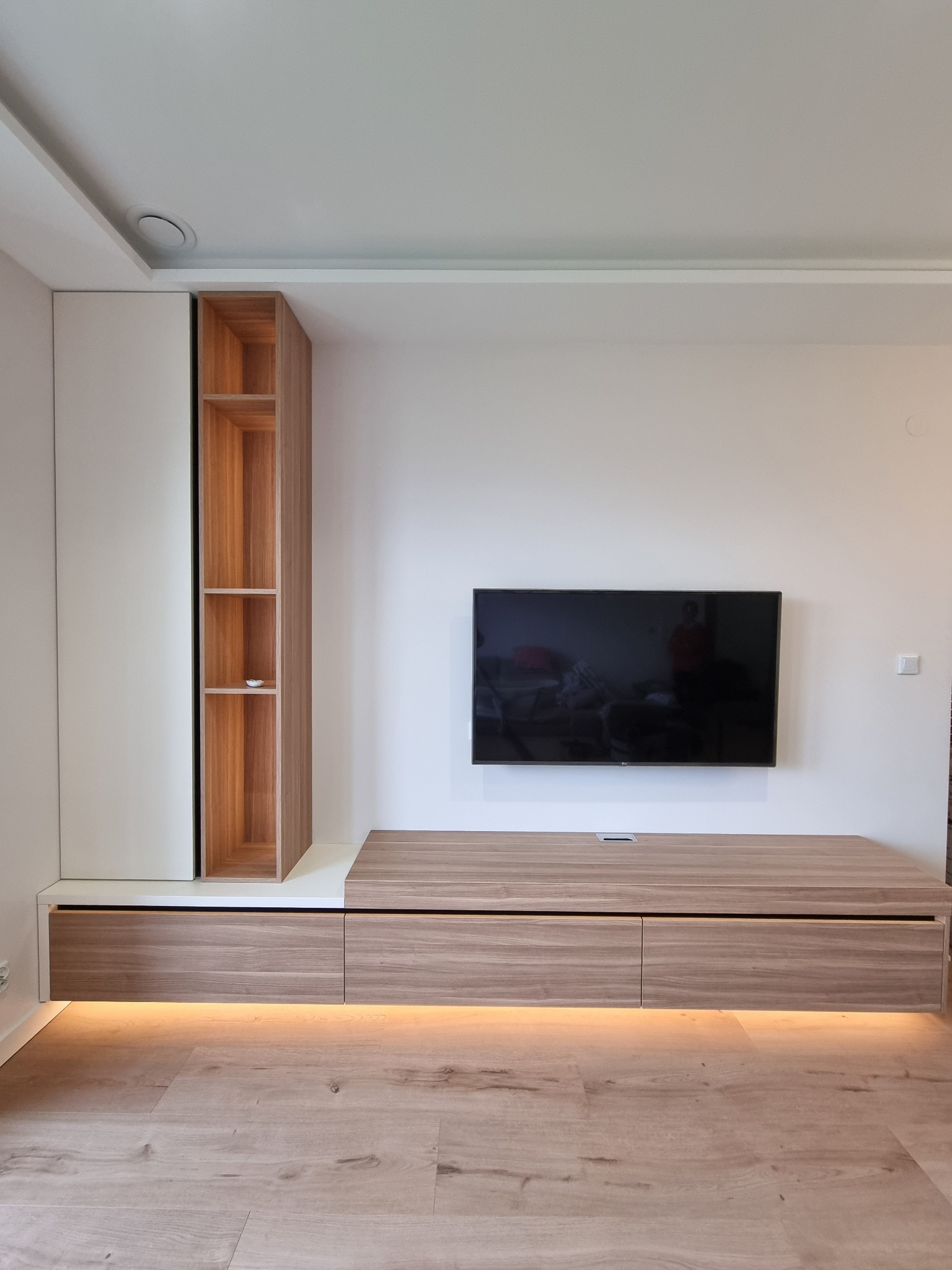 Nowoczesna zabudowa ściany telewizyjnej z białymi szafkami, drewnianym regałem z półkami i podświetlanym szafkami RTV, na podłodze panele drewnopodobne.